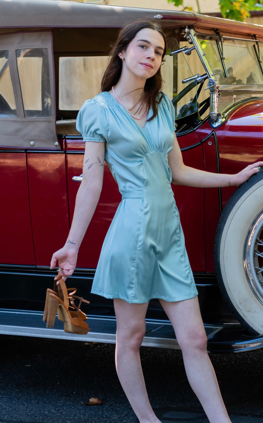 A female model wearing a lime green mini dress with short sleeves. Posing against a vintage car background.