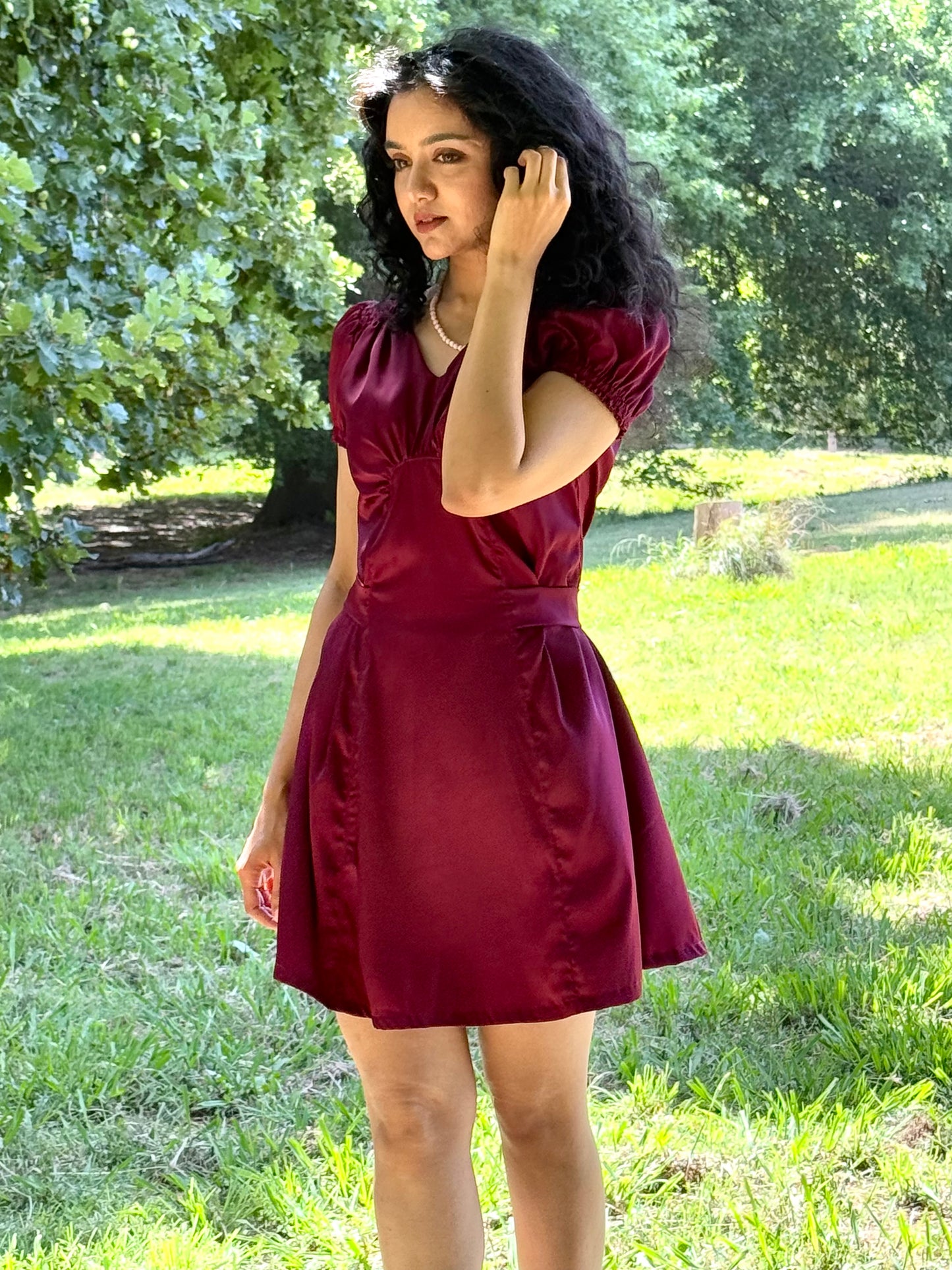 A female model wearing a wine red mini dress, posing against a garden background.